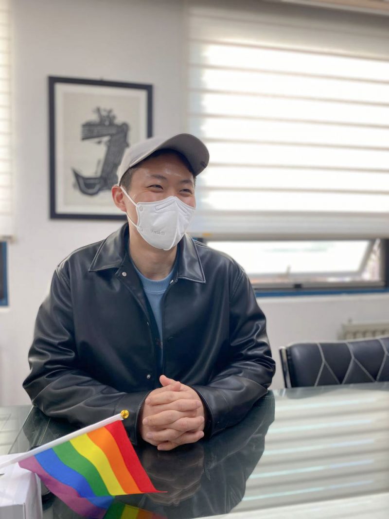 장길완 활동가가 유리테이블 위에 깍지 낀 손을 얹은 채로 의자에 앉아 있다. 회색 모자와 흰색 마스크, 가죽 점퍼를 입은 그의 눈에 웃음기가 있다. 그의 앞으로는 프라이드 손깃발이 놓여있다.
