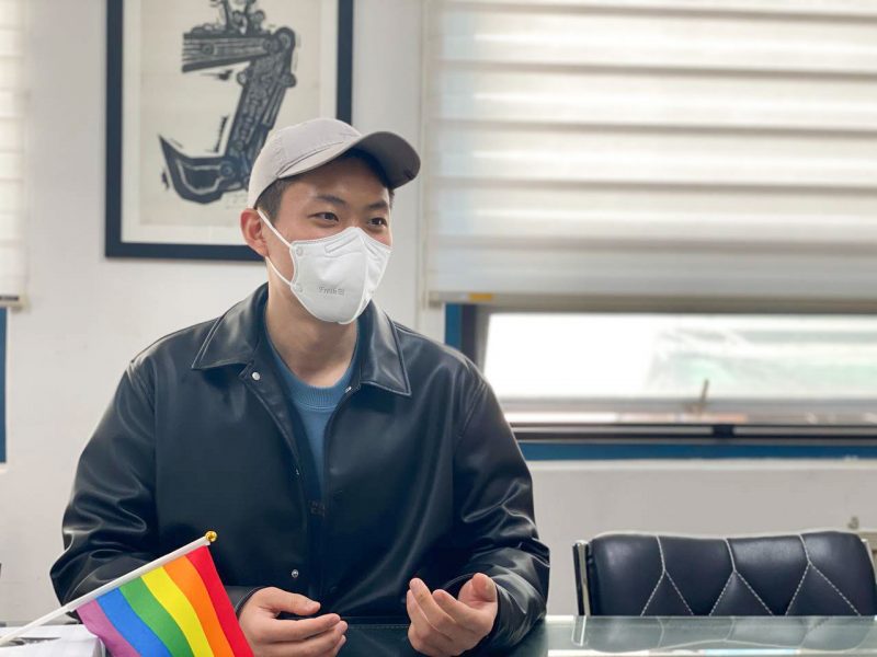 장길완 활동가가 유리테이블 위에 두 손을 손바닥이 위로 가게 얹은 채로 의자에 앉아 있다. 회색 모자와 흰색 마스크, 가죽 점퍼를 입고 있는 그의 앞으로는 프라이드 손깃발이 놓여있다.